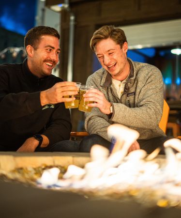 Two men smiling and clinking their beer glasses in front of a firepit